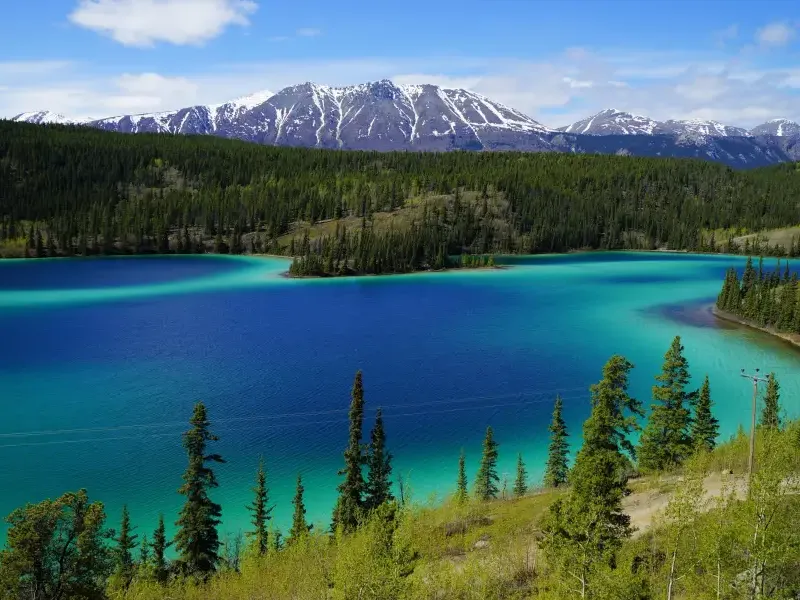Vivid turquoise and deep blue glacial lake surrounded by boreal forest and snow-capped mountains in Alaska wilderness.