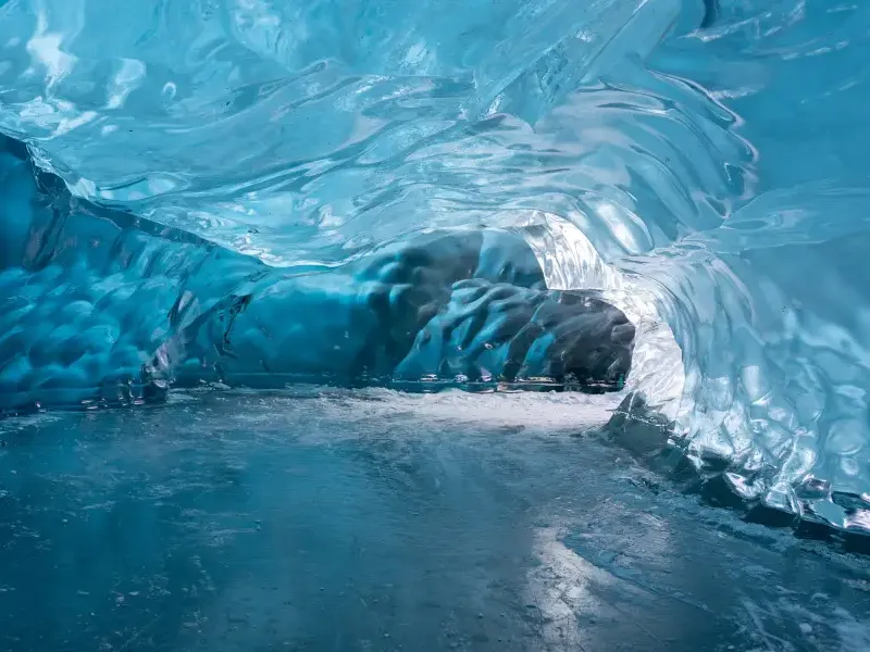 Glowing blue ice cave tunnel inside an Alaska glacier; with smooth frozen walls and icy floor leading to a dark passage.