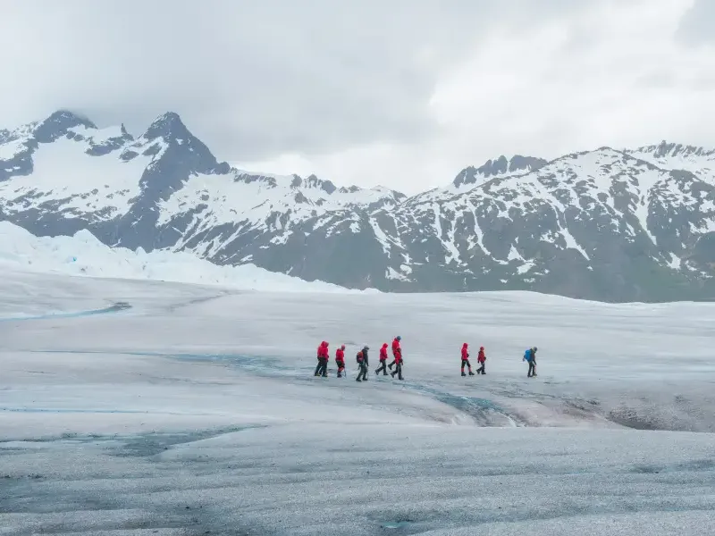 Group of hikers in red jackets trekking across an Alaska glacier with snow-capped mountains in the background.
