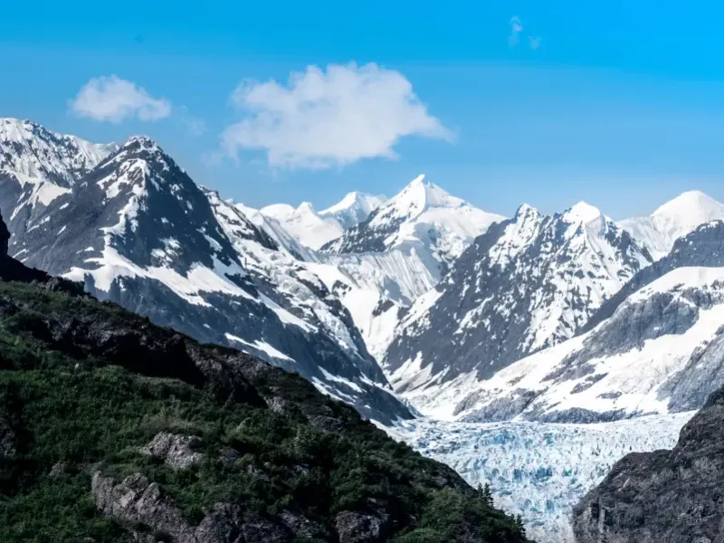 Snow-capped mountains frame a flowing glacier through a valley in Alaska under a clear blue sky.