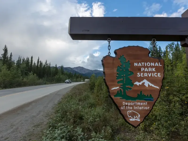 National Park Service arrowhead sign hanging roadside in Alaska; with forested highway and mountain range in background.