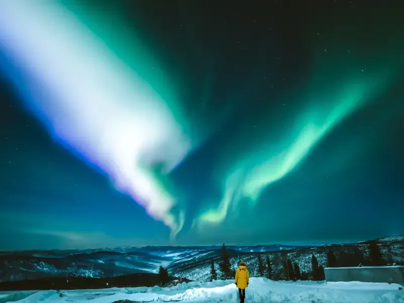 Traveler in yellow coat gazing at vivid green and white aurora borealis over snow-covered Alaska mountains at night.
