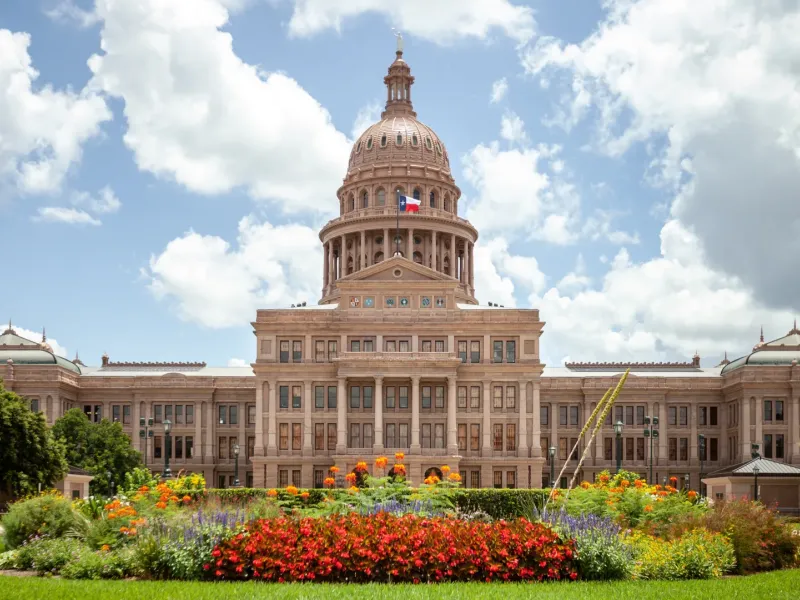 Austin Texas State Capitol building with copper dome Texas flag and colorful flower gardens under a partly cloudy sky