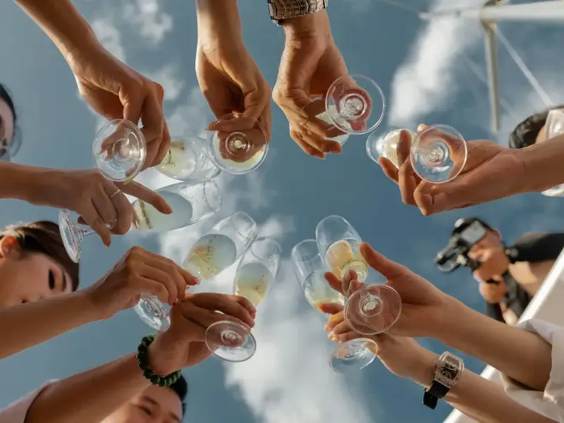 Group of guests raising champagne glasses in a circle toast on a cruise; photographed from below against a blue sky.