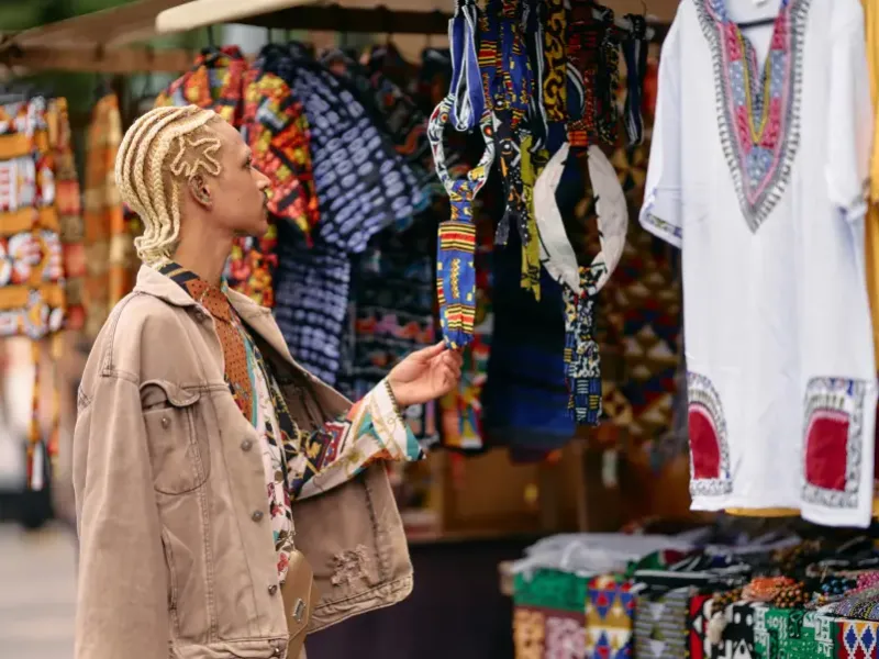 Traveler with blonde braids browsing colorful African textiles at an outdoor market stall during a cruise-land excursion.
