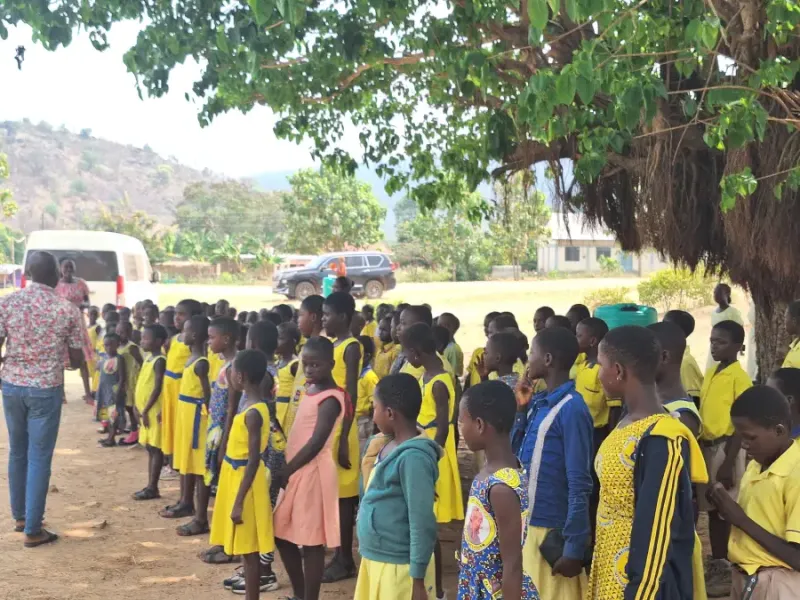 Students in yellow uniforms lined up outdoors under a tree during eat-mea-2024 school assembly with a teacher present.