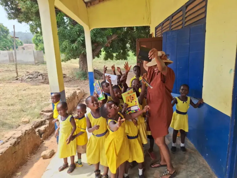 Adult in straw hat takes selfie with smiling children in yellow uniforms outside a school during EAT-MEA-2024 visit.