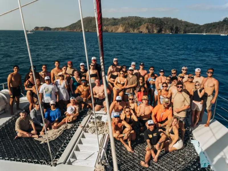 Large group of travelers posing together on a catamaran cruise deck with tropical island coastline in the background.