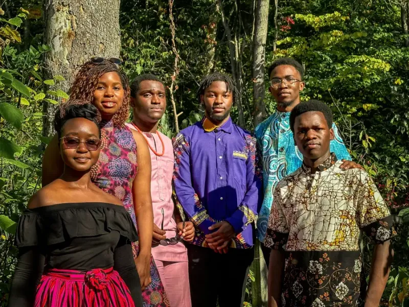 Six adults in vibrant African print attire standing together among sunlit green trees.