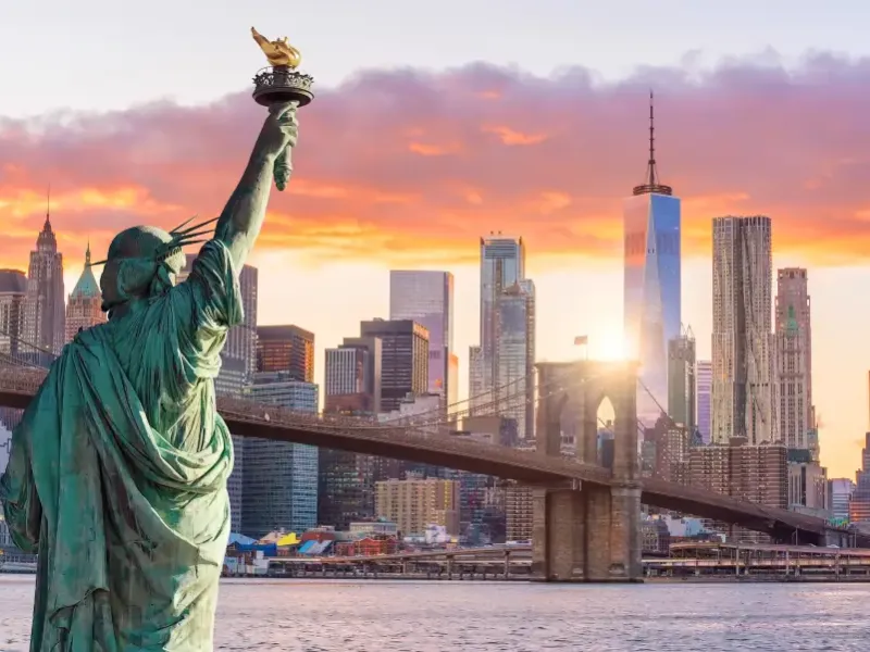 Statue of Liberty faces the New York New York skyline at sunset with Brooklyn Bridge and One World Trade Center behind