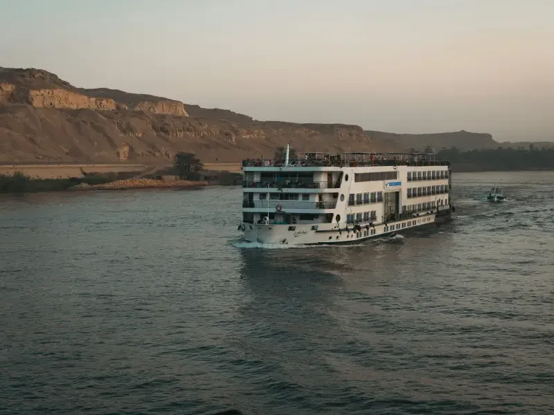 Multi-deck river cruise ship sailing the Nile at dusk with desert cliffs and palm trees in the background.