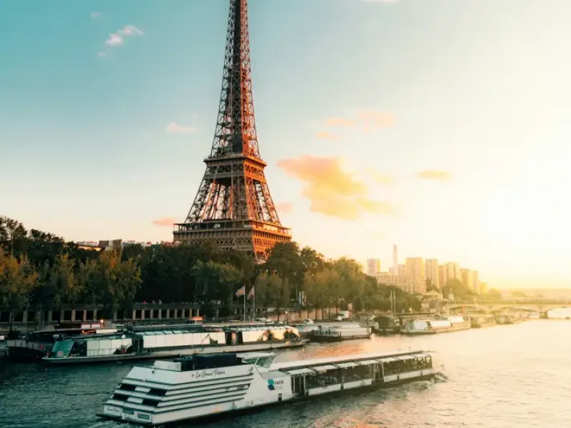 Paris Seine River cruise boat passing the Eiffel Tower at golden sunset; with city skyline in background.