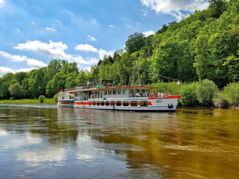 White and red river cruise vessel sailing along a calm tree-lined river under a partly cloudy summer sky.