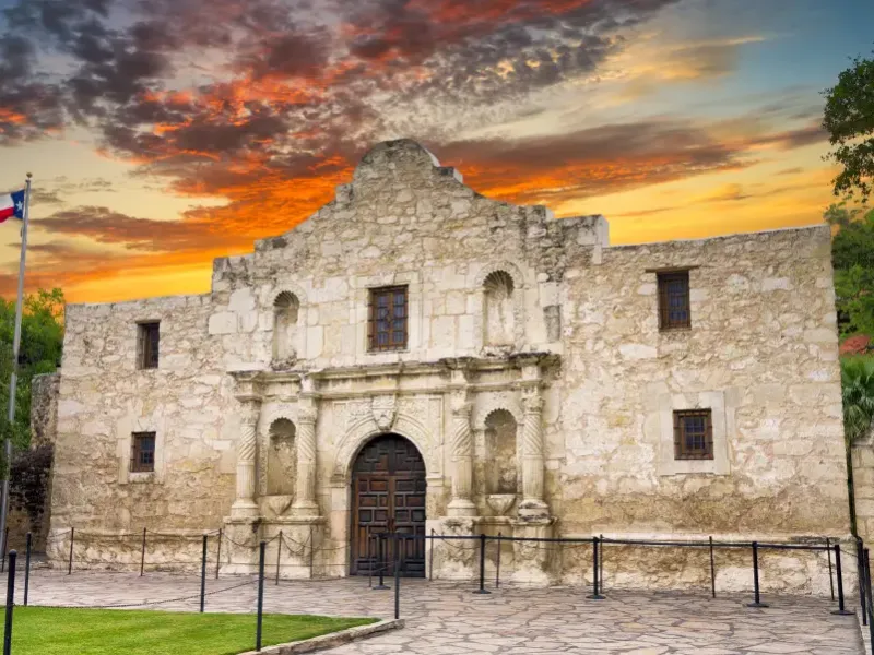 The Alamo's limestone facade at sunset in San Antonio Texas with the Texas flag flying and a dramatic orange sky