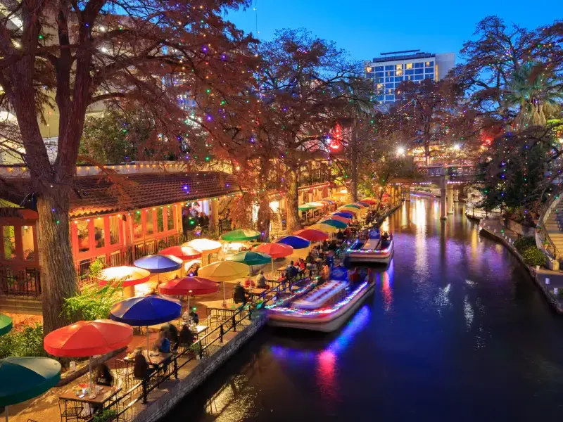 San Antonio Texas River Walk at dusk with colorful holiday lights umbrella-lined restaurants and a lit barge gliding through the canal