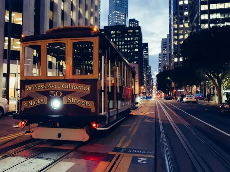 San Francisco CA cable car number 50 on California Street at dusk; with downtown skyscrapers and city lights in background.