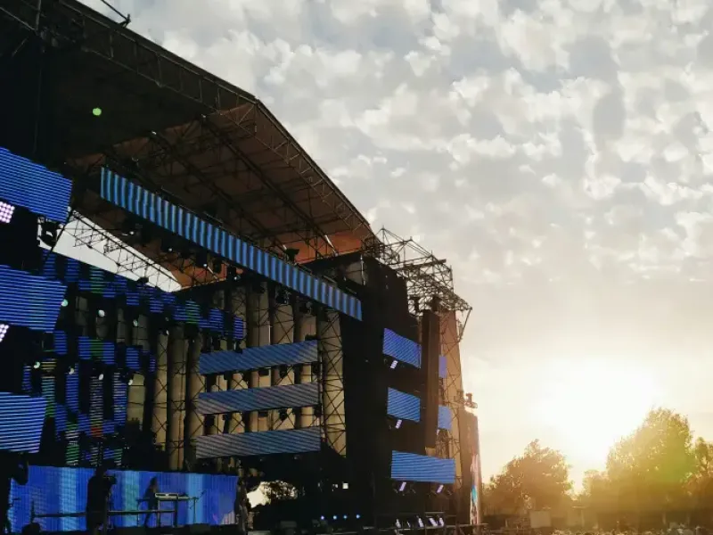 Large crowd at a Chicago outdoor music festival at sunset; with LED stage screens and a rainbow pride flag visible.