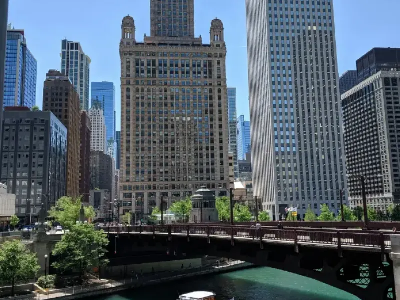 Packed tour boat on the turquoise Chicago River passes under a bridge with the Jewelers Building and Kemper tower beyond.