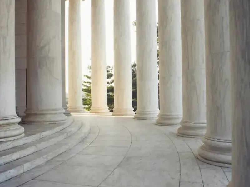 Curved marble colonnade of the Jefferson Memorial in Washington; DC; with Ionic columns framing sunlit trees beyond.