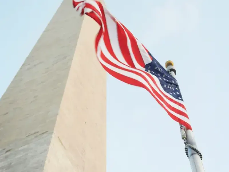 American flag waving beside the Washington DC Monument obelisk viewed from below against a clear blue sky.
