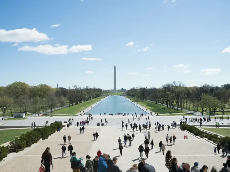Tourists walk along the National Mall in Washington DC with the Reflecting Pool and Washington Monument in the background.