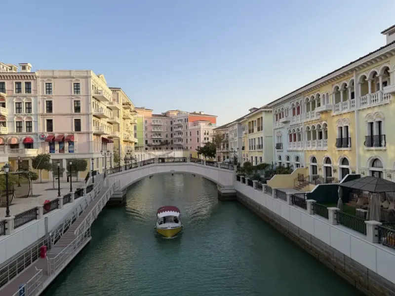 A yellow water taxi moves along a canal lined with pastel Venetian-style buildings and a pedestrian bridge in Doha Qatar.