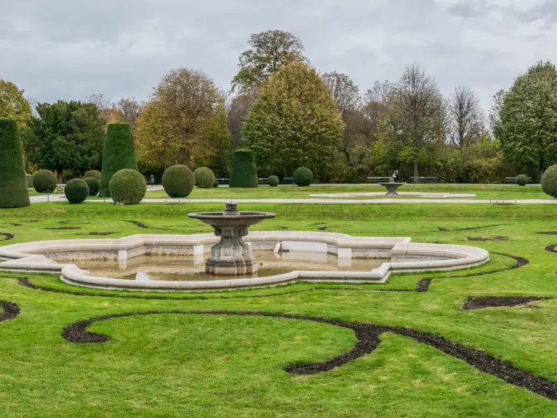 A clipped parterre and fountain in the gardens of Schoenbrunn Palace in Vienna under a gray and white sky.