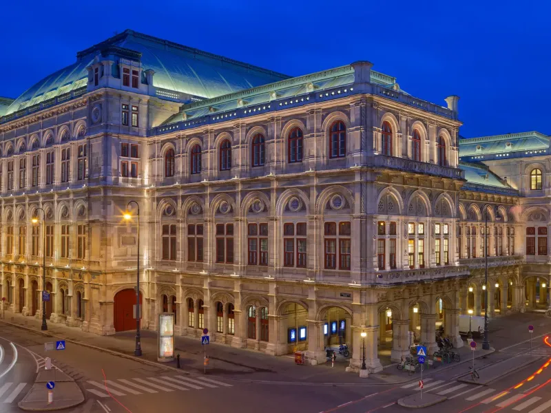 The lit facade of the Vienna State Opera at blue hour, with the wet carriageway of the Ringstrasse in front of it.