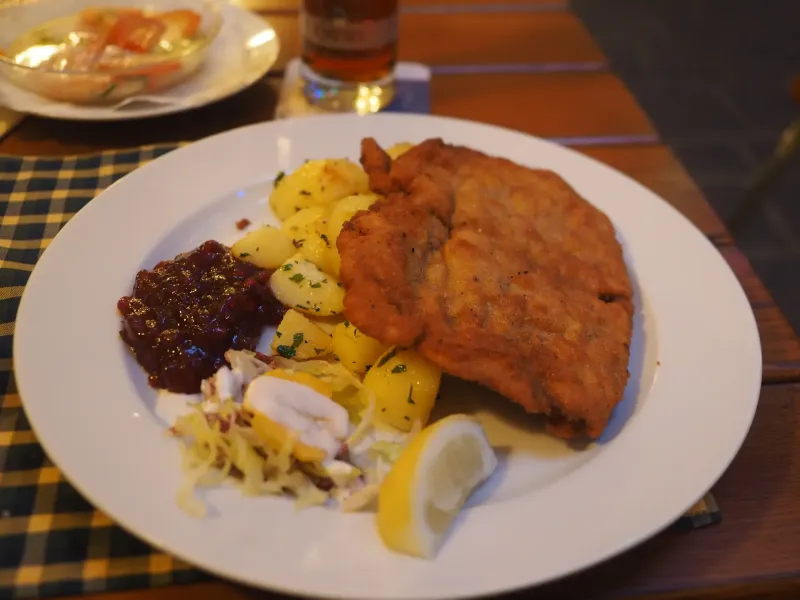 A Wiener Schnitzel served on a white plate with parsley potatoes, lingonberry preserve and a wedge of lemon.
