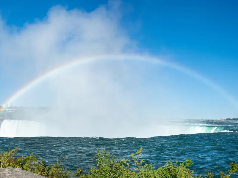 The curved crest of Horseshoe Falls throwing spray high above the gorge, with a full rainbow arcing across the mist.