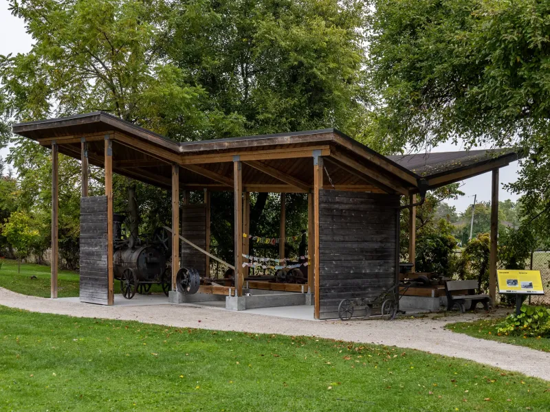 A timber shelter housing a steam engine and sawmill bench, with a bilingual interpretive panel beside the gravel path.