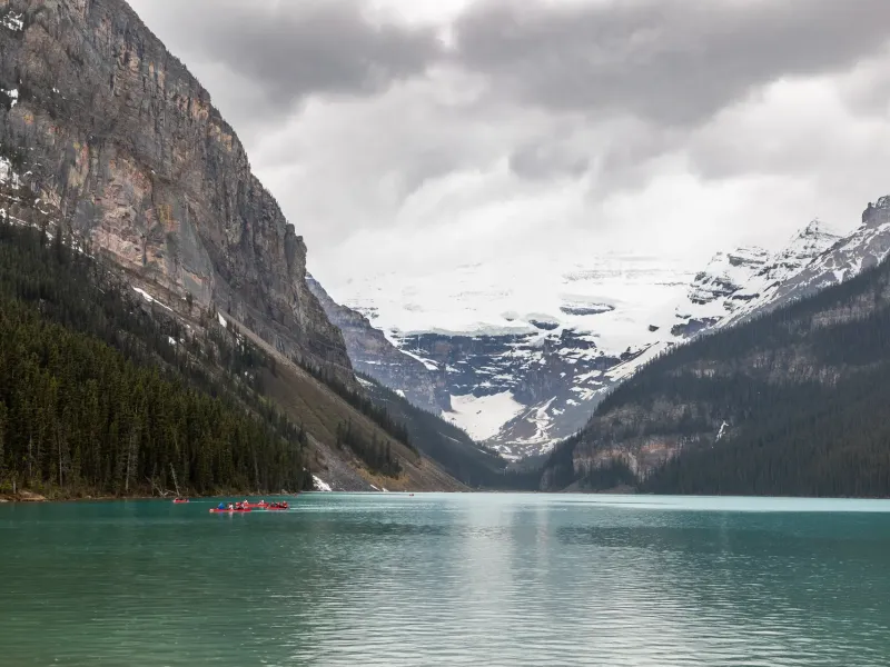 A pale green lake between forested cliffs, a snow and ice field filling the valley head, and red canoes near the shore.