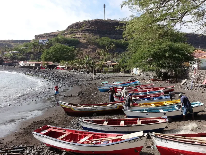 Painted open boats drawn up on the dark beach at Cidade Velha, with the fortified ridge above.