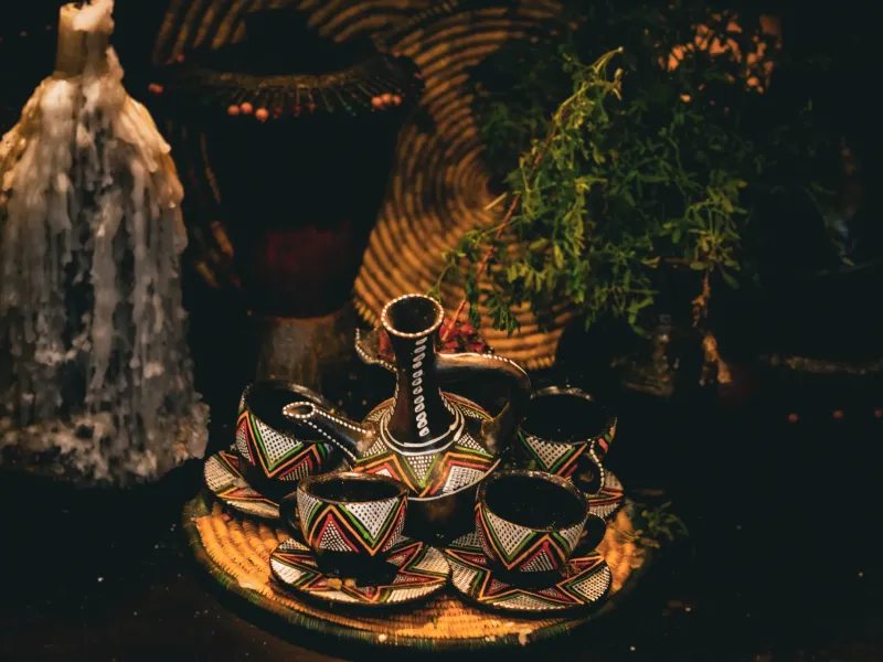 A clay jebena and small handleless cups on a decorated tray for a coffee ceremony, with grass strewn around them.