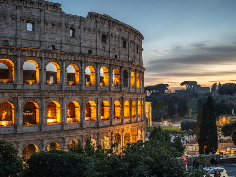 The Colosseum in Rome at dusk, its arcades lit from within, with the ruins of the Roman Forum beyond