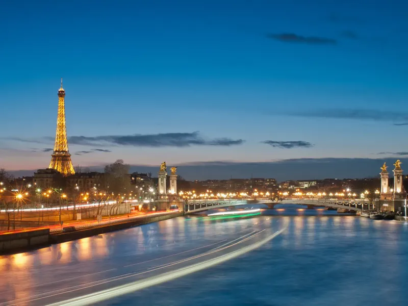 The Eiffel Tower lit at dusk above the Seine in Paris, with the gilded columns of the Pont Alexandre III to the right