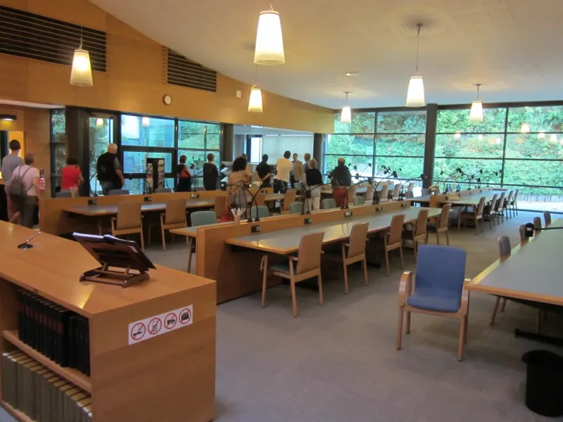 Long reading tables with individual lamps and bound registers on a shelf in a French departmental archives room.