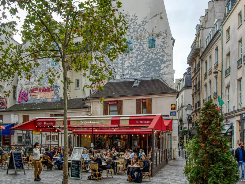 A full cafe terrace under red awnings on a small Paris square, with tables close together and the street beyond.