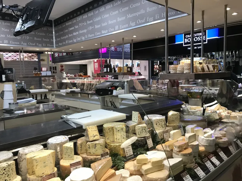 Wheels and rounds of cheese massed across a covered market counter under a hand lettered board in Lyon, France.