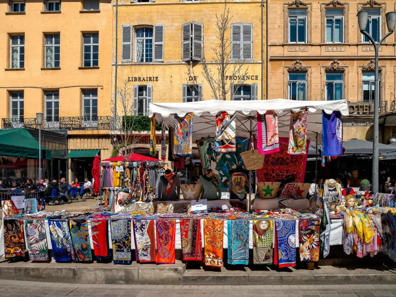 A market stall hung with printed scarves and textiles under a white awning on a shaded square in Provence, France.