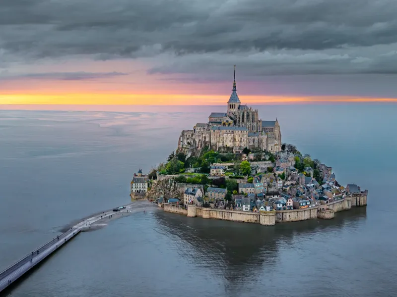 Mont-Saint-Michel rising from the bay at dusk with the modern causeway running out to it across the shallow water.