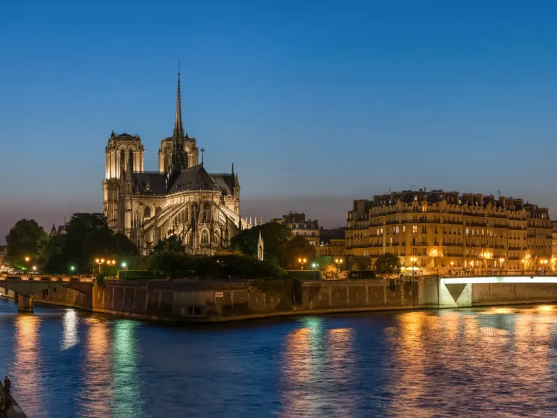 The spire and flank of Notre Dame above the lit quays of the Ile de la Cite, reflected in the Seine at dusk in Paris.