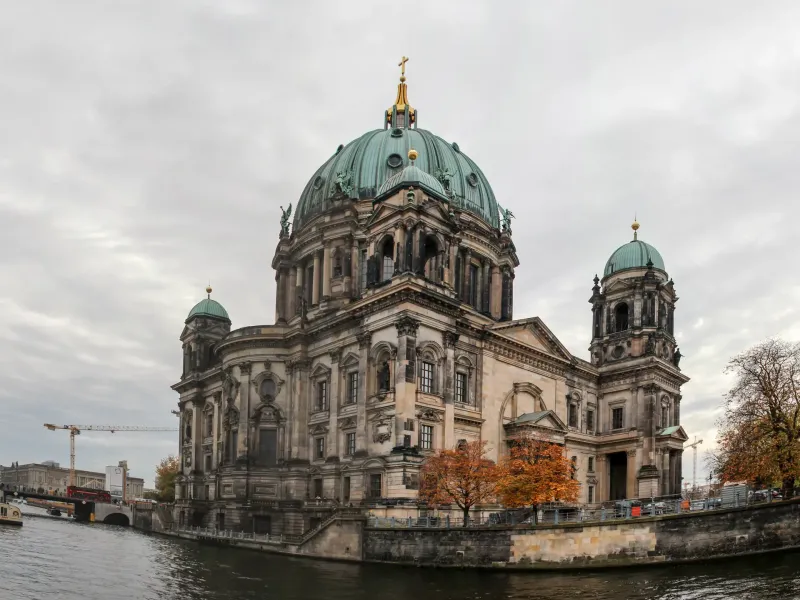 The green dome of Berlin Cathedral above the Spree embankment, with autumn trees along the water in front of it.