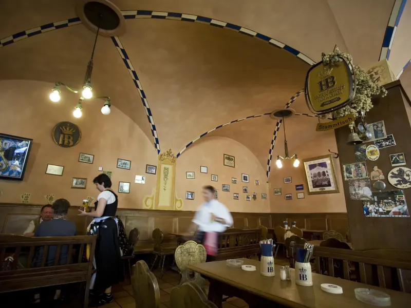 The vaulted interior of a Munich beer hall with long shared tables, a server carrying steins and the walls hung with prints.