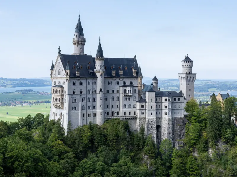 Neuschwanstein Castle standing on its wooded spur in daylight, with the lakes and flat farmland of the Bavarian plain behind.