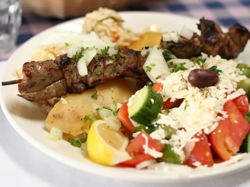 A plate of lamb souvlaki with potatoes, tzatziki and a Greek salad of tomato, cucumber and grated cheese.