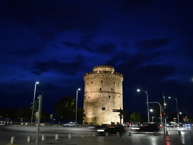 The floodlit White Tower of Thessaloniki against a deep blue evening sky, with traffic passing in front.
