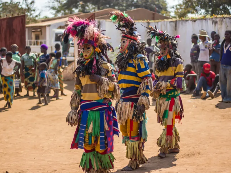 Masked Gule Wamkulu dancers in tall feathered headdresses and fringed costumes on open ground, a crowd watching.