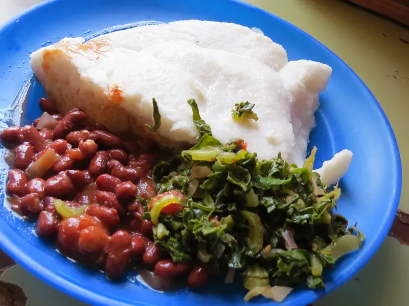 A blue enamel plate holding two portions of white nsima with stewed beans, cooked greens and a piece of fish.