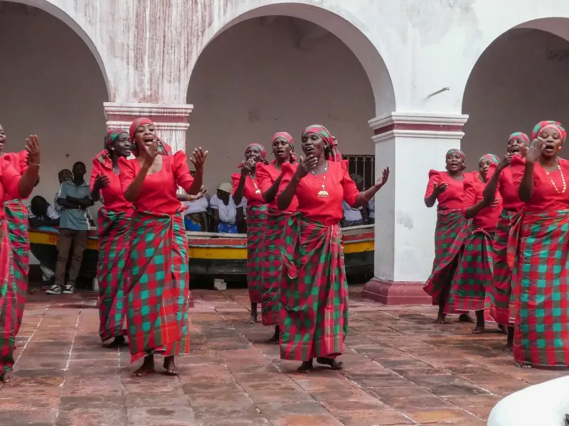 Women in matching red tops and checked capulana wraps singing and clapping in an arcaded courtyard in Mozambique.